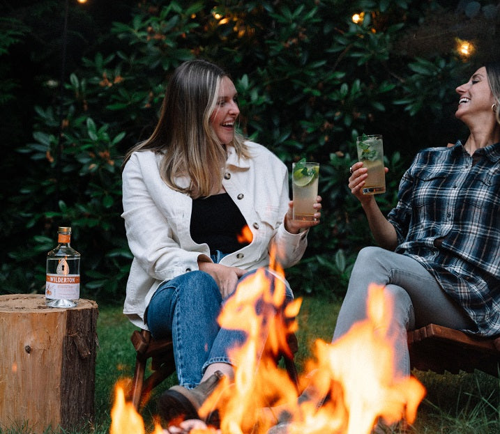 two women sitting around a campfire holding drinks