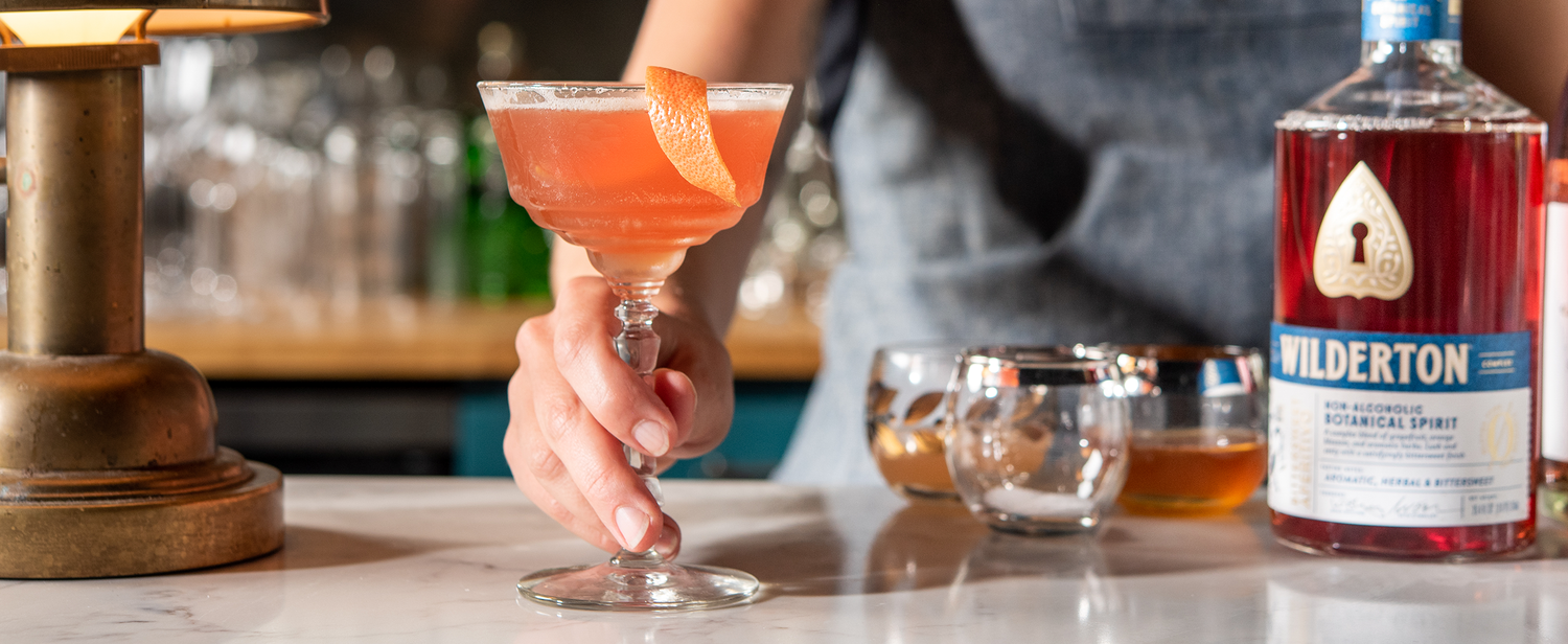 hand around the stem of a glass with an orangish pink drink on a bar next to a bottle of bittersweet aperitivo