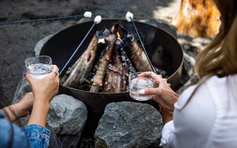women's hands holding glasses with clear drinks around a campfire
